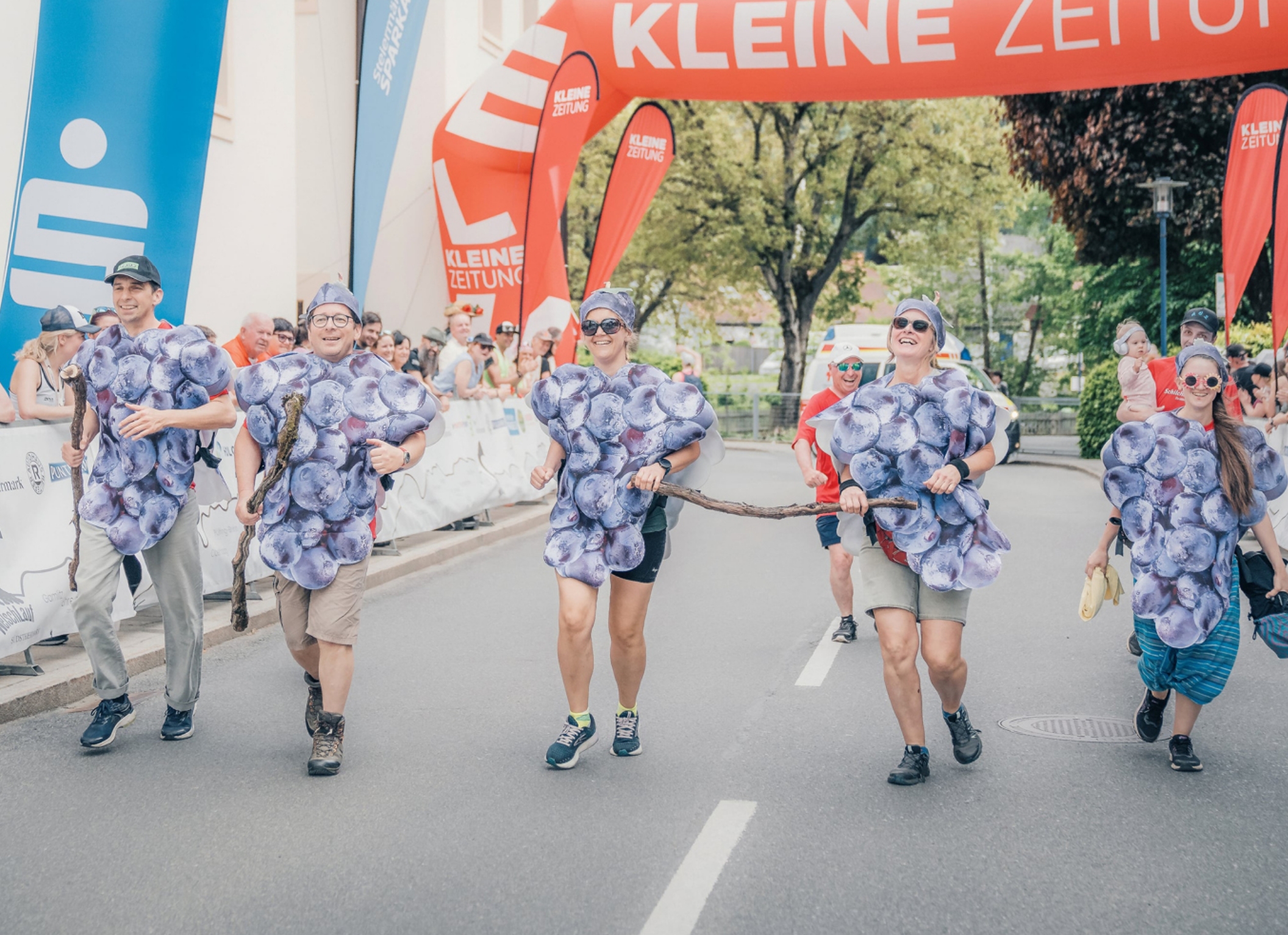 5 Personen als Weintrauben verkleidet laufen zusammen in das Ziel, am Streckenrand stehen zahlreiche Zuschauer. Das Kostüm besteht aus einer Kappe mit einem Weintraubenblatt und einen Weintrauben-Poncho der über die Sportbekleidung gezogen ist. Die Stimmung ist lustig, locker und spaßig.
