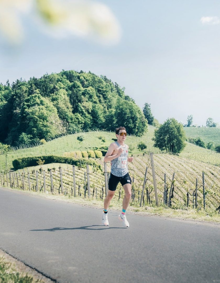 welsch-marathon-vorschau Ein Mann läuft in Sportbekleidung auf einer Straße, umgeben von grünen Weinbergen und Wald. Sonnenschein mit sommerlichen Temperaturen.